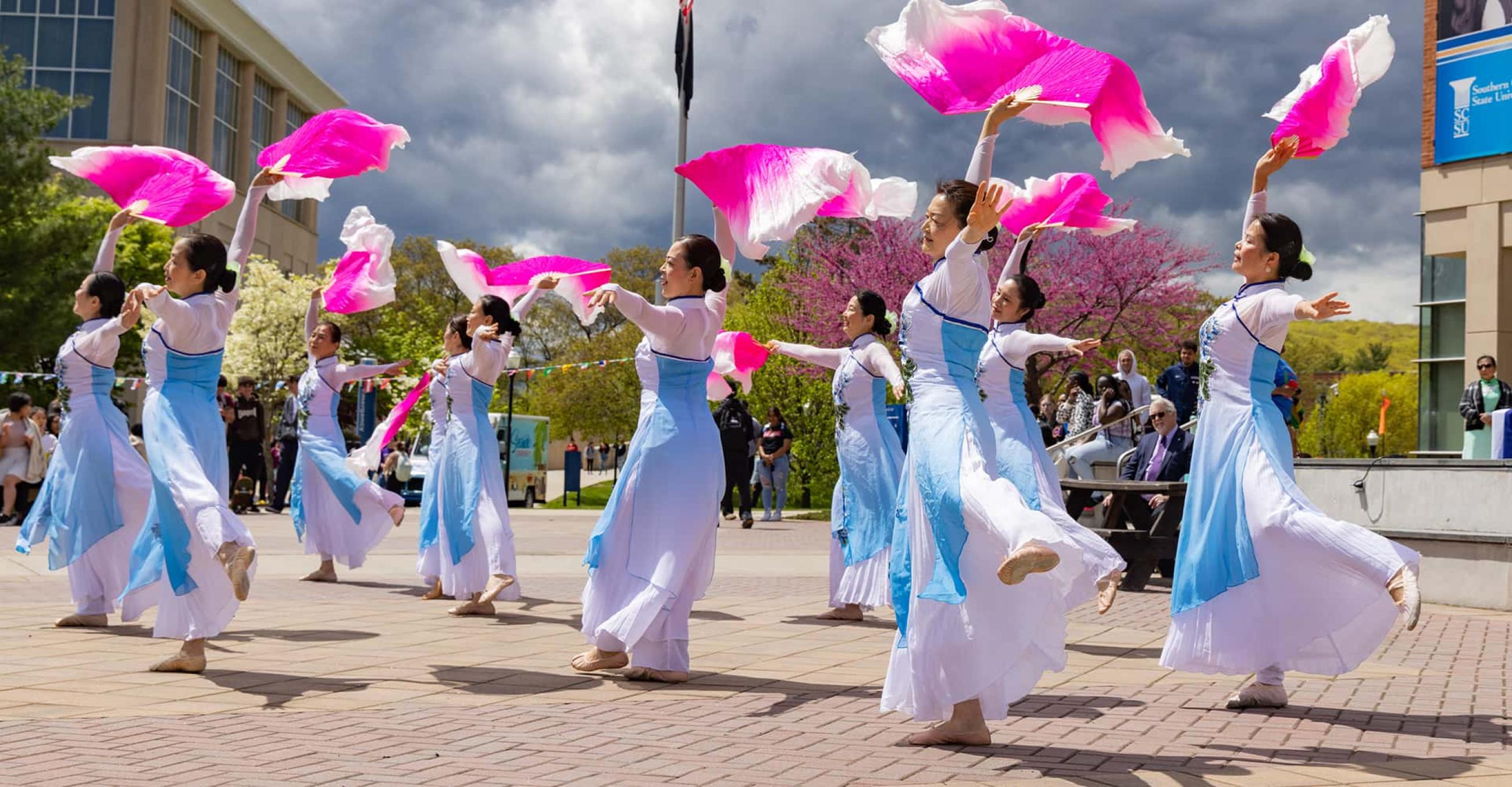 A group of dancers in traditional Asian costume dance with pink fans outdoors on Buley Patio