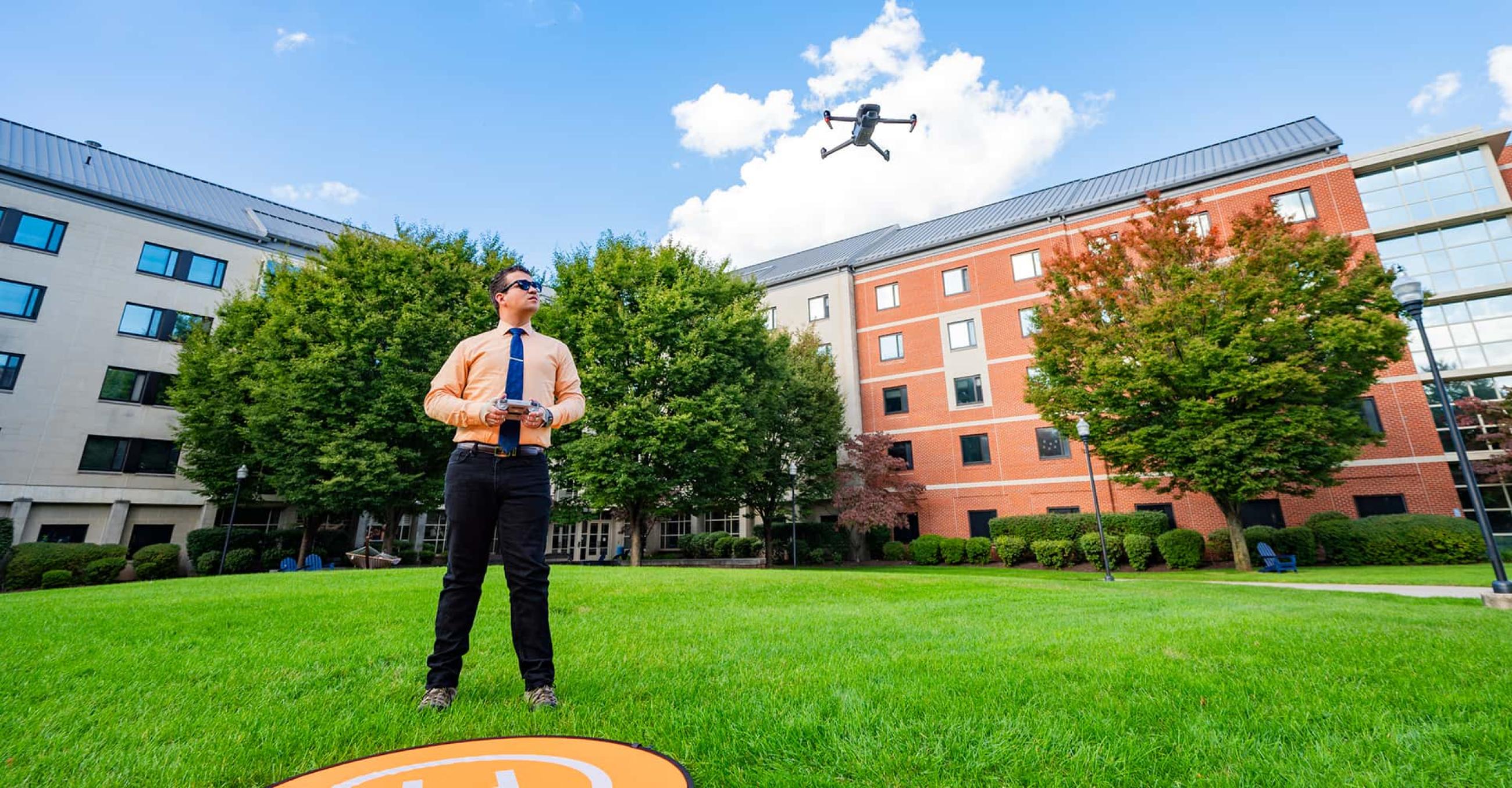 a man stands holding a controller and a drone flies just above him