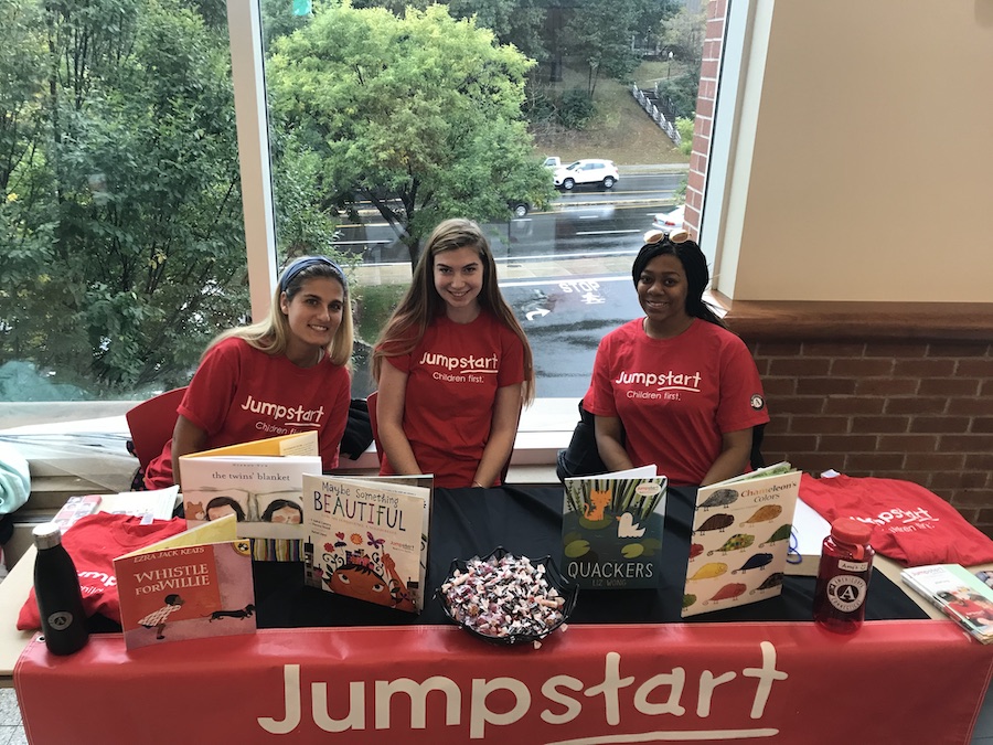 Students wearing red Jumpstart program shirts sitting around a table of books and program promotion giveaways