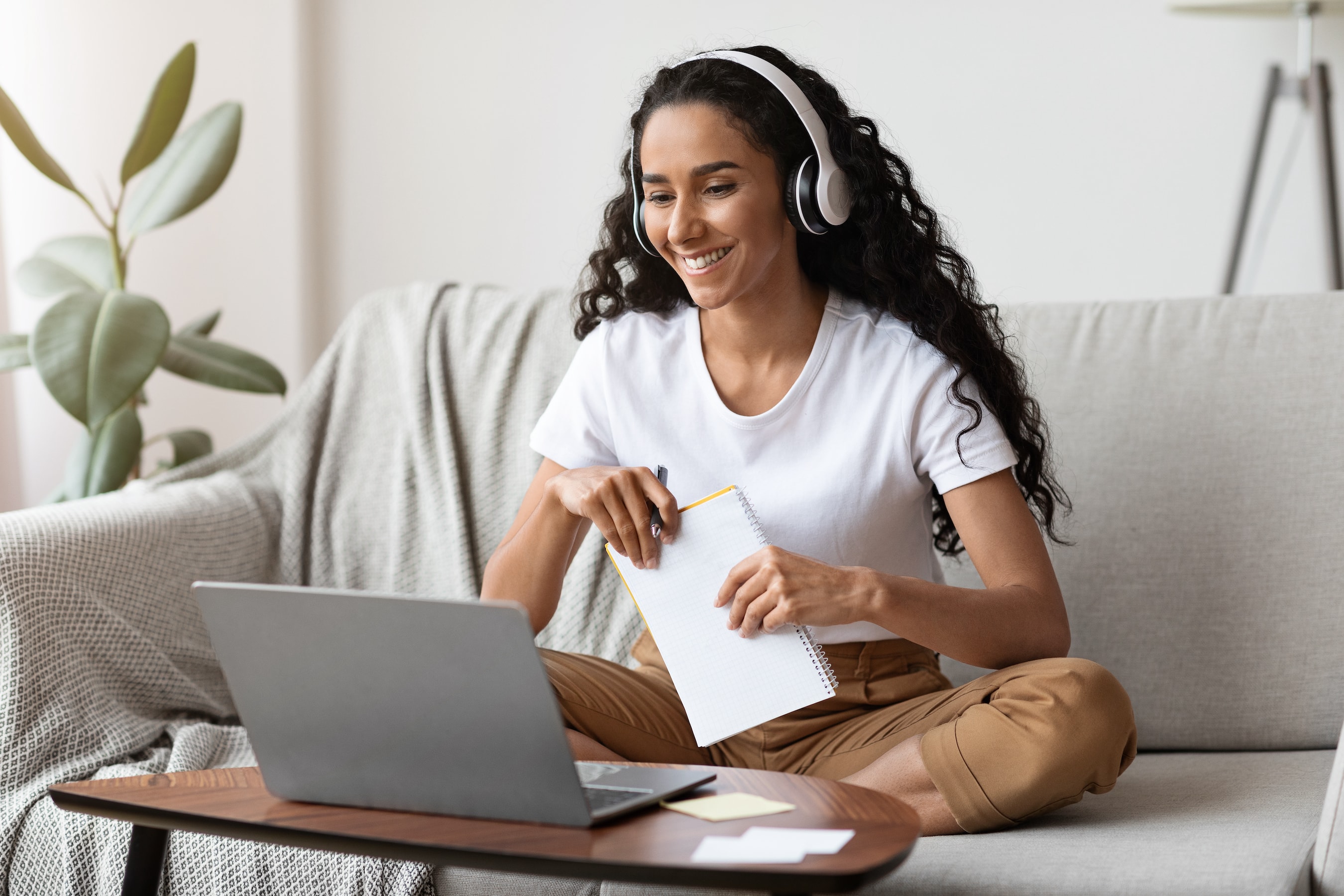 "A woman with headphones on, sitting at home on a couch, taking online classes"