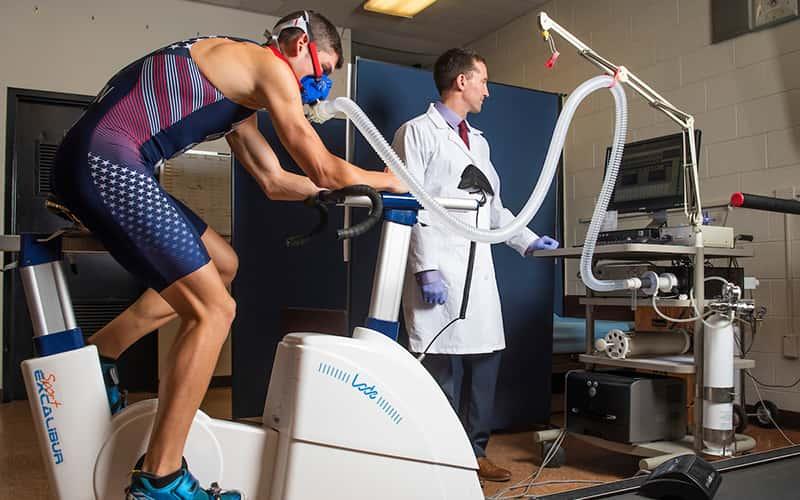 a male riding a stationary bike is monitored by a professor in a white coat