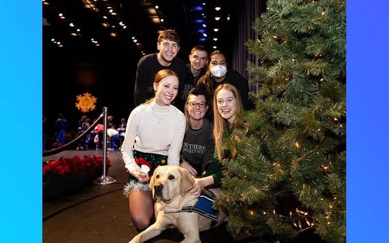 a group of students sit with a yellow Labrador Retriever beside a Christmas tree