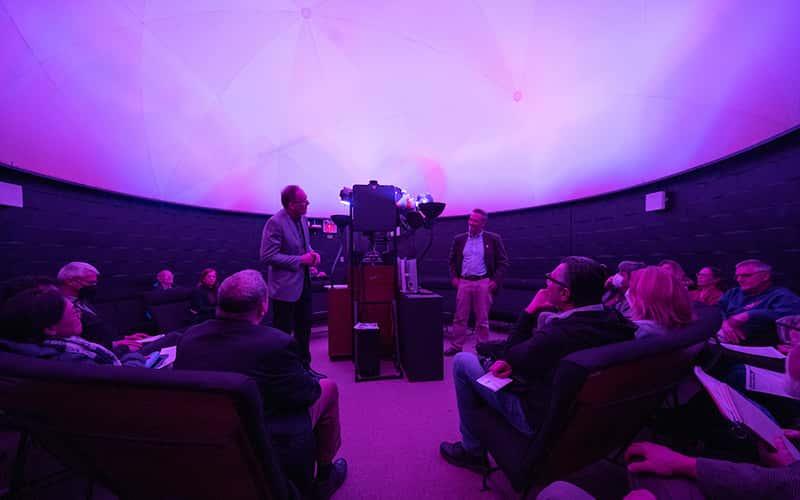 two professors stand in front of an audience, next to a projector inside the campus planetarium