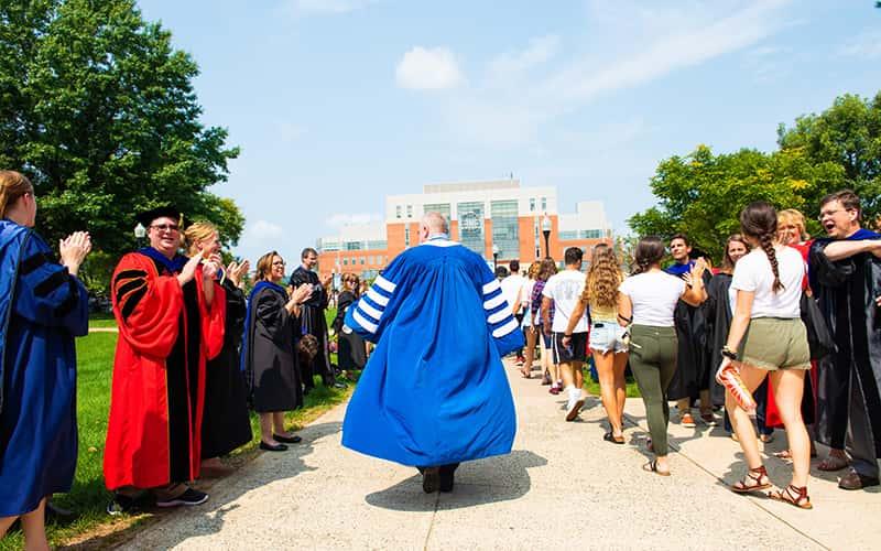 president bertolino walks away from the camera with his blue academic robe flowing behind him