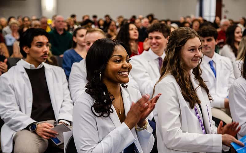 graduating nursing students in white coats sit in the audience at their pinning ceremony