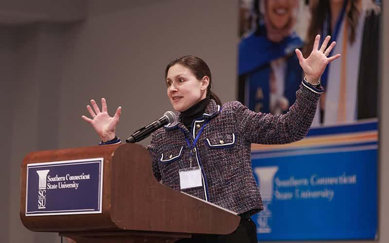 a speaker stands behind a podium with her arms outstretched, apparently speaking animatedly