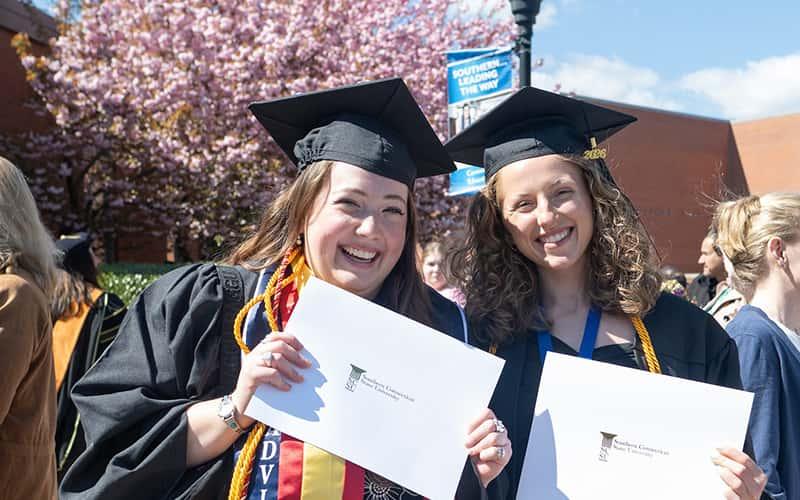 Two smiling students in graduation caps and gowns hold up white certificate folders