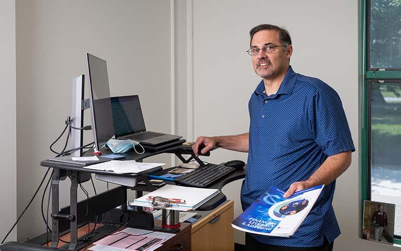 Lew DeLuca stands at his computer, holding brochures about financial literacy