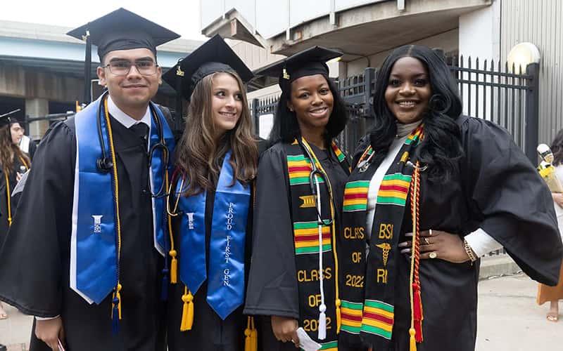 four students in graduation caps and gowns; one wears a blue stole that says "Proud First-Gen"