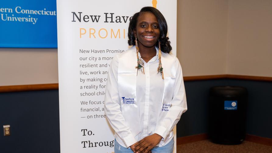 A female student in front of the New Haven Promise sign with a UAP Graduation Sash.