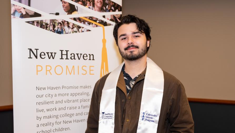 A male student in front of the New Haven Promise sign with a UAP Graduation Sash.