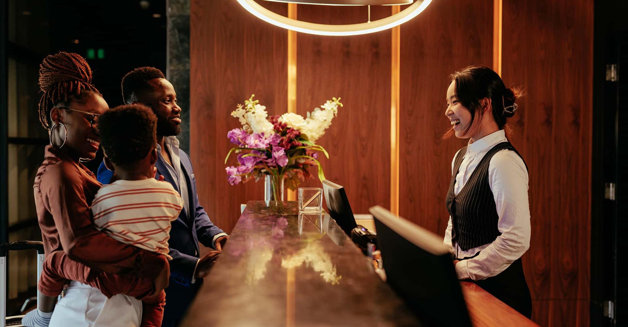 a woman behind a hotel reservation desk greets a man and woman with a small child
