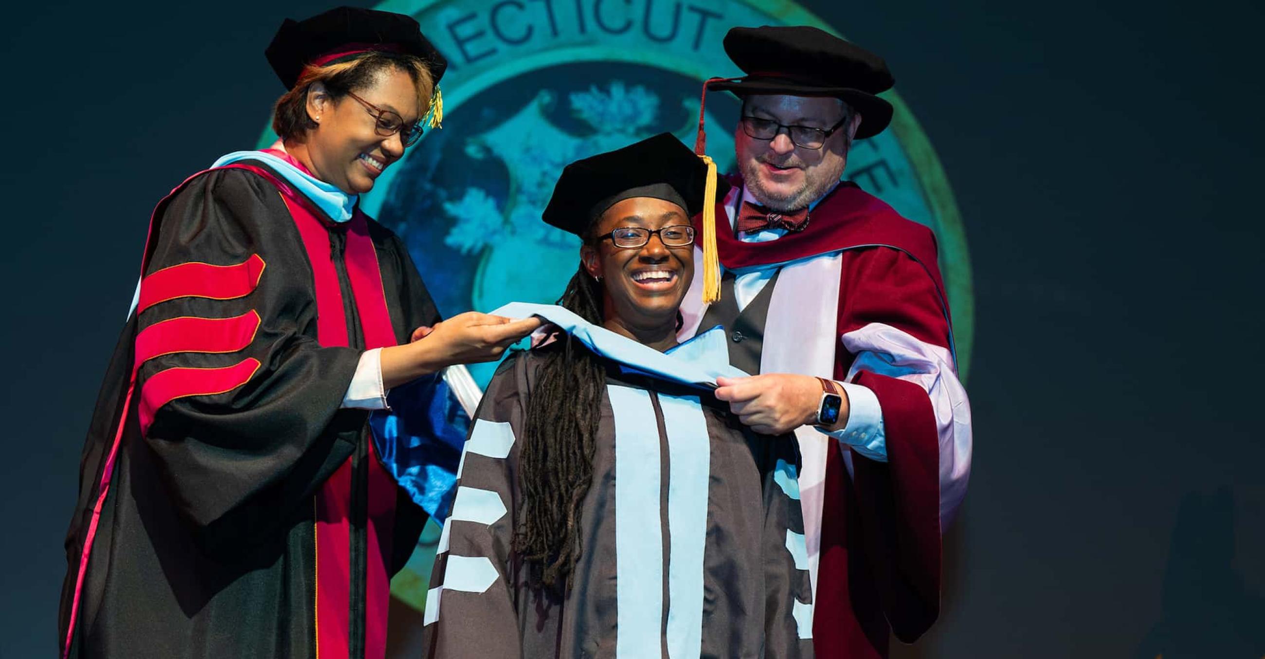 a student at commencement receives her hood from two faculty members