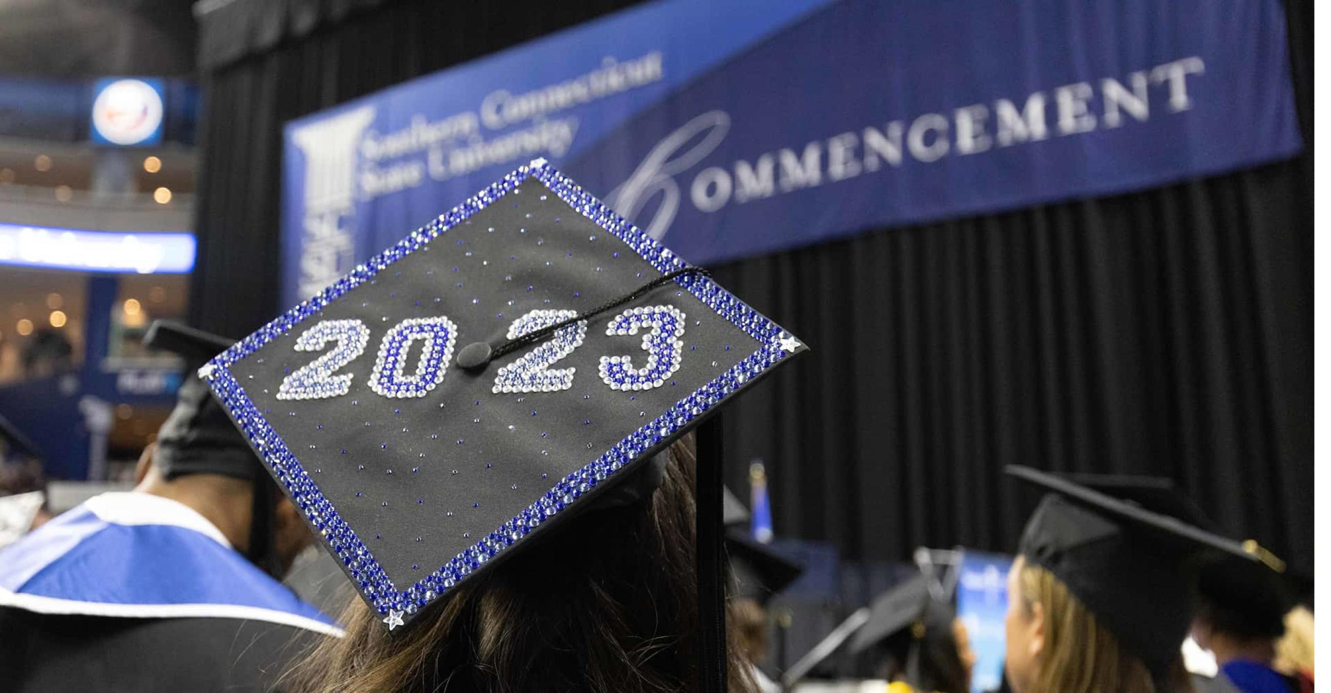 a student's graduation cap with "2023" printed on it, with the undergraduate commencement ceremony in the background