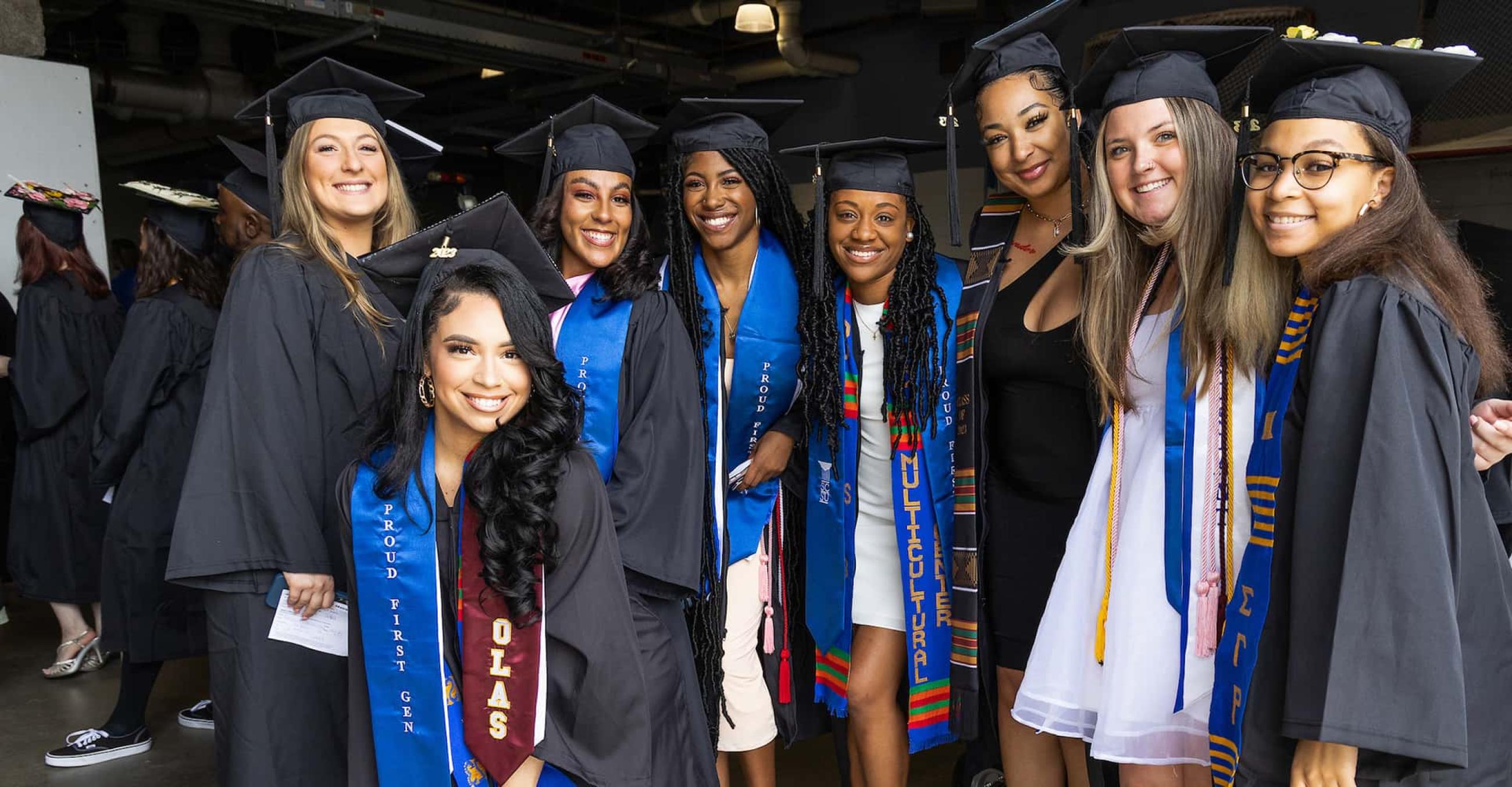 a group of smiling students in caps and gowns at Commencement, all with "Proud First-Gen" stoles