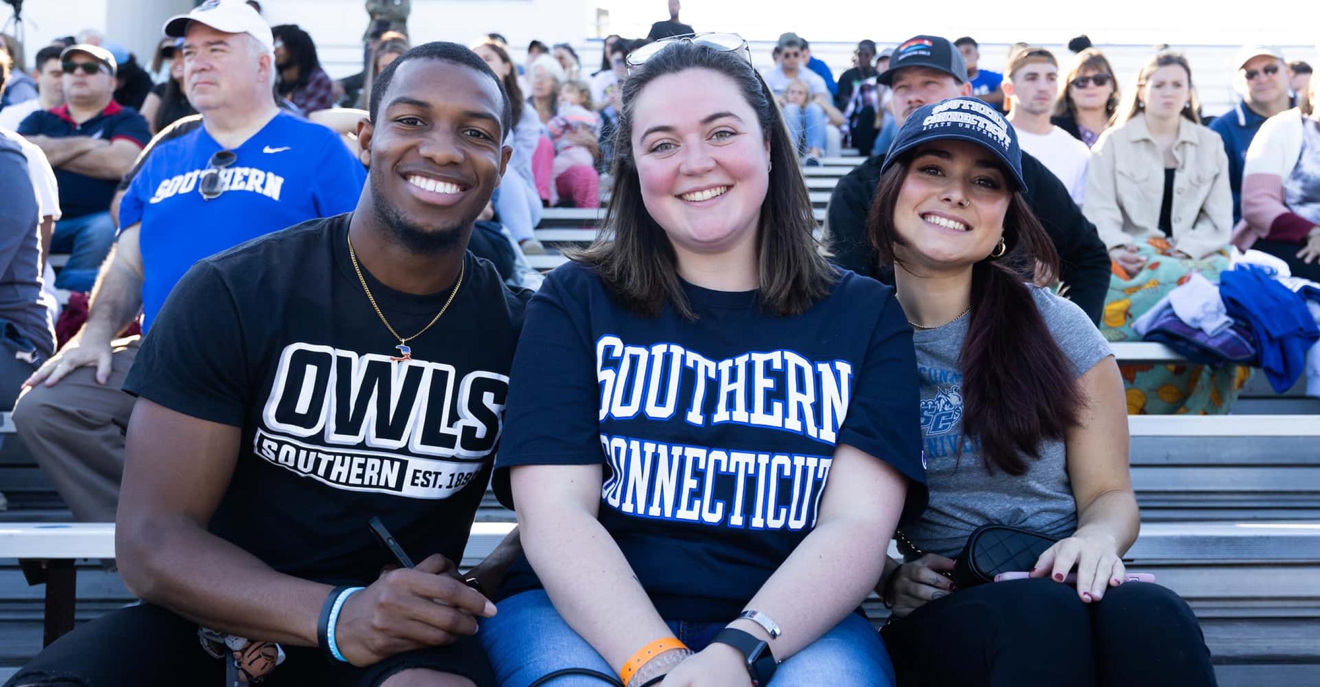 three students sit in the football stands and smile at the camera