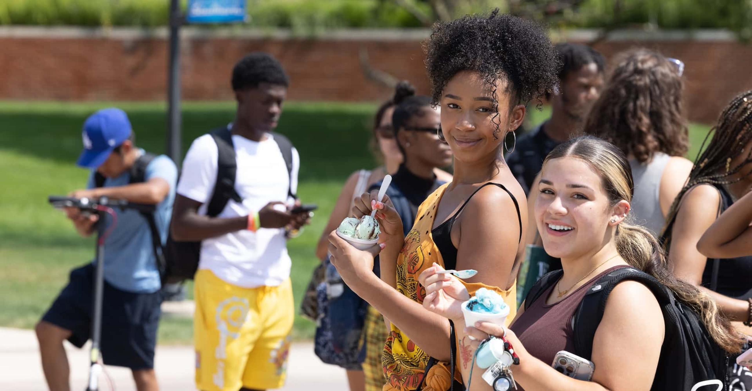 two students eating ice cream smile for the camera while other students mill about in the background