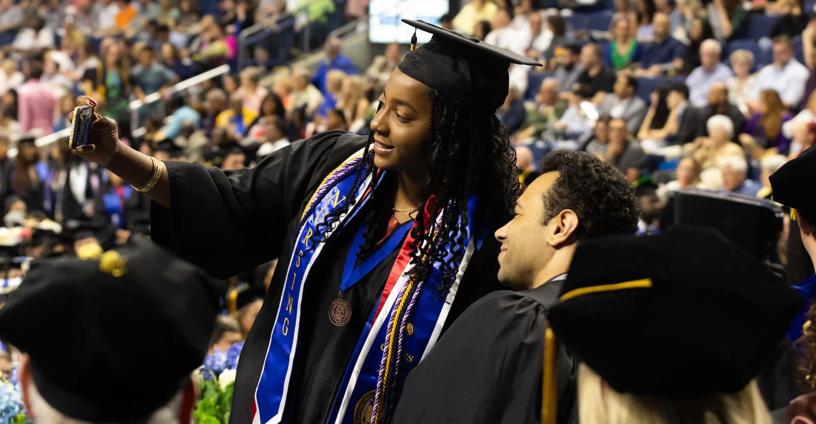 a student in cap and gown pauses to take a selfie with Commencement speaker Corbin Bleu