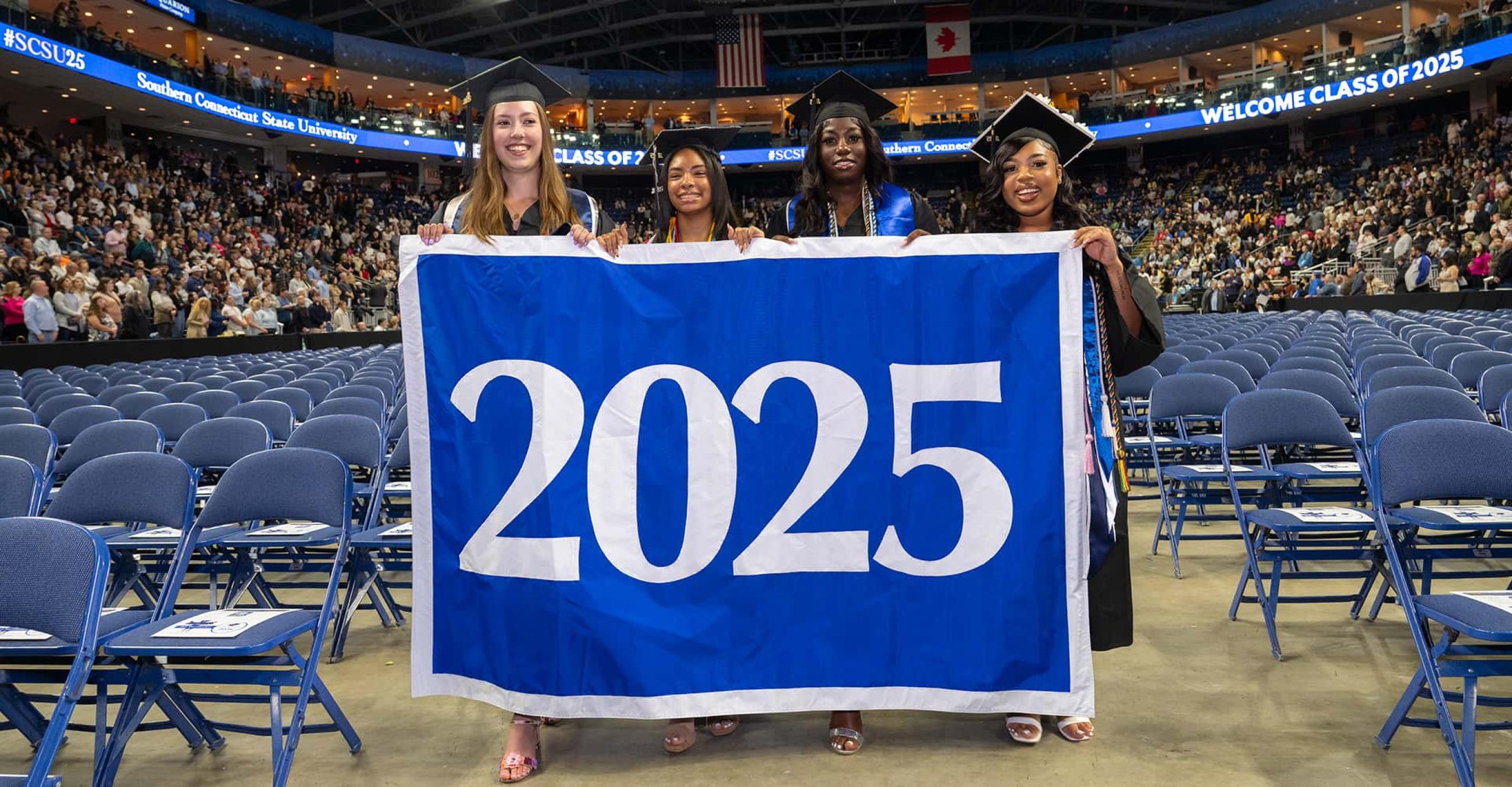 four students in graduation caps and gowns carry a banner that says in large numerals "2025"