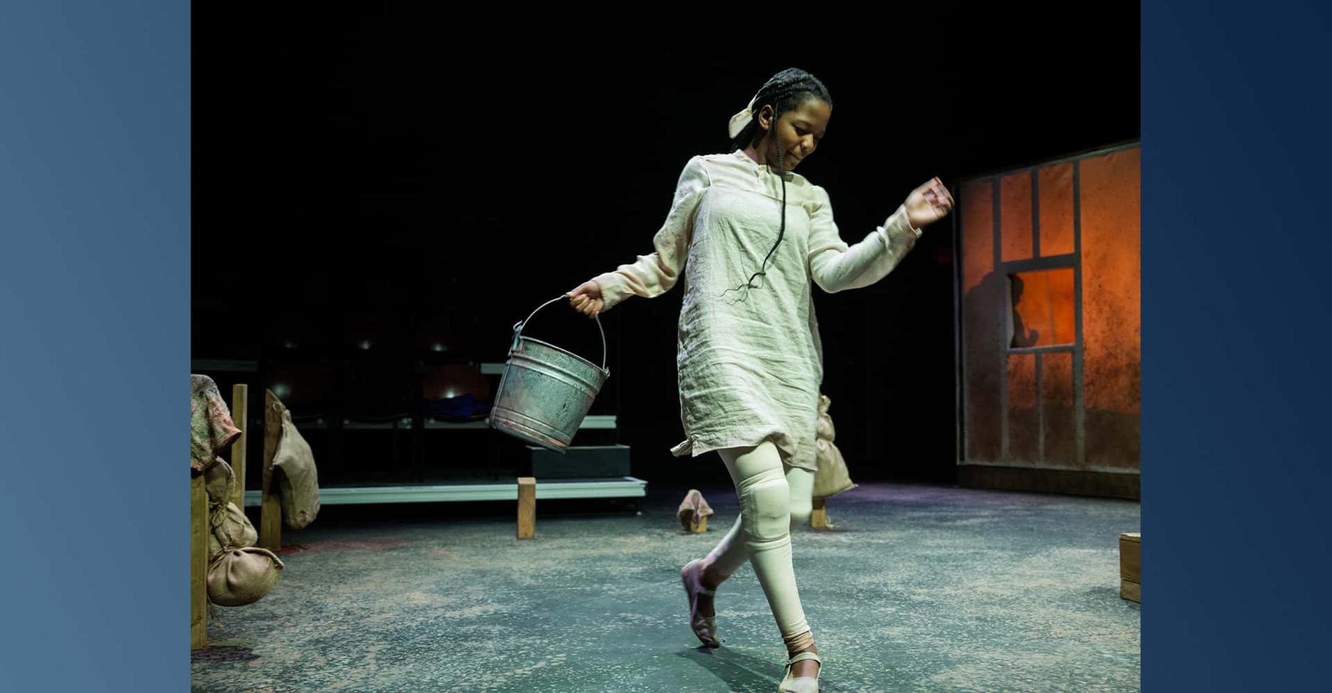 a young woman carrying a metal bucket appears to dance on a stage