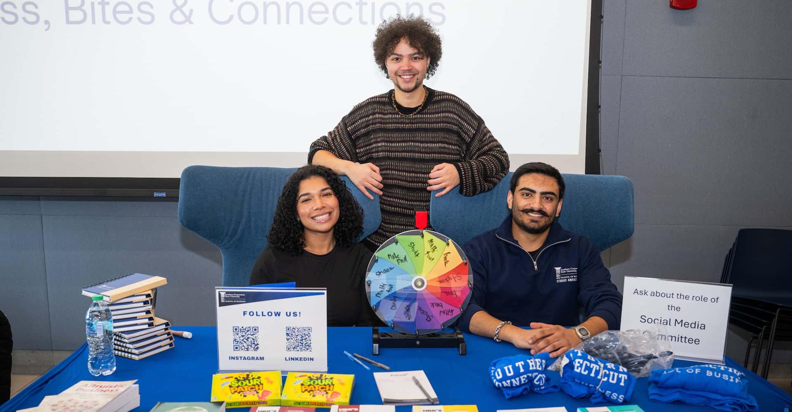 three smiling students sit behind a table with materials promoting a club