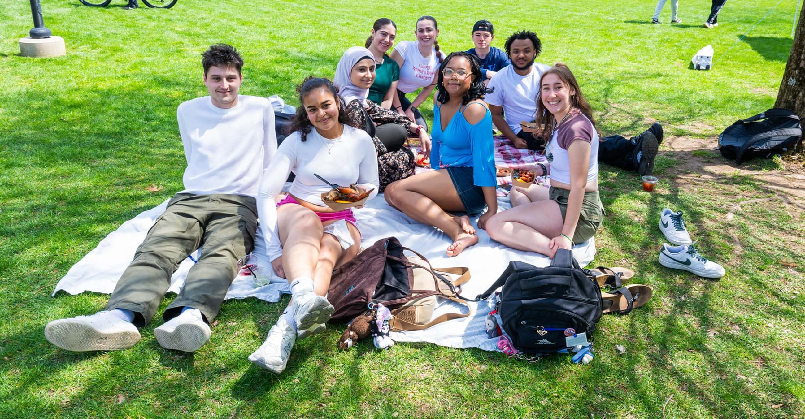 a group of students sit on a blanket on the academic quad, smiling at the camera