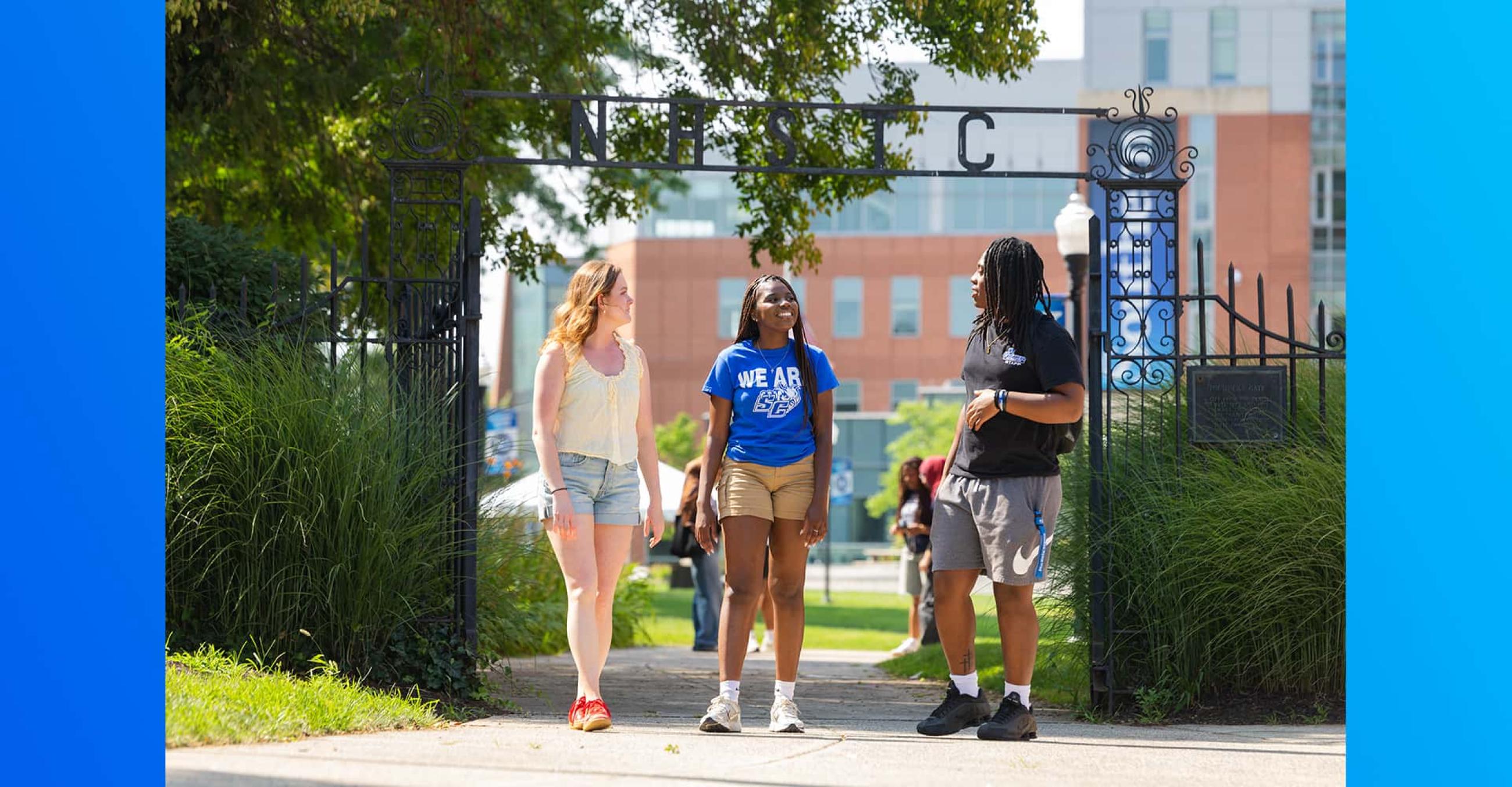three students stand, chatting, in front of Founders Gate on the SCSU campus